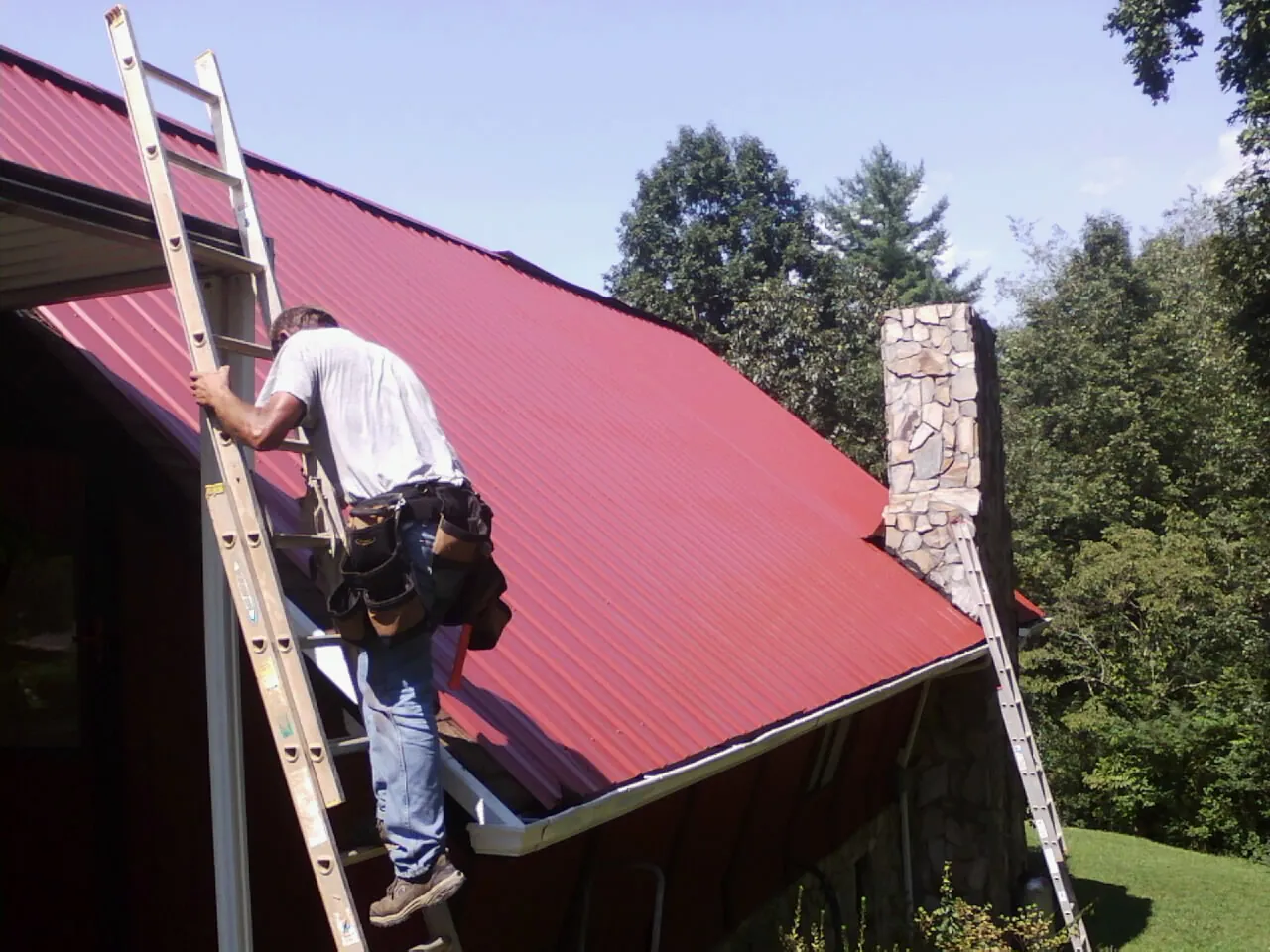 Roofer on a ladder beside a red metal roof and stone chimney — Soffit & Fascia Repair by Roofing Brothers in Whittier